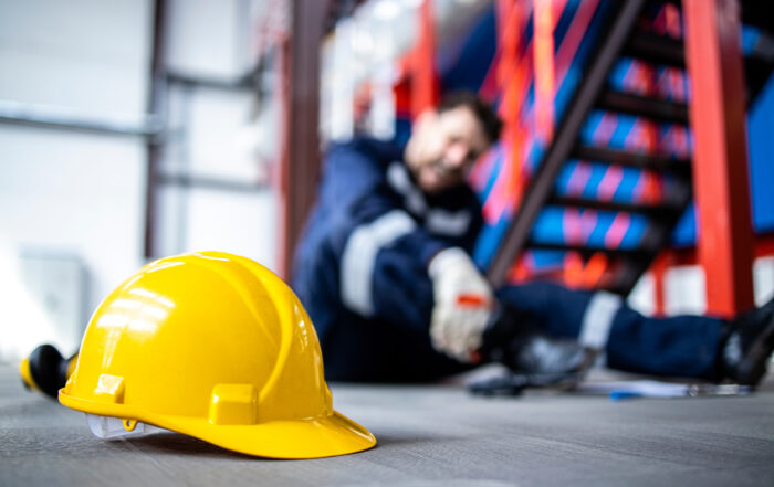 Yellow safety helmet on the floor with an injured worker holding his leg in the background of an industrial workplace.